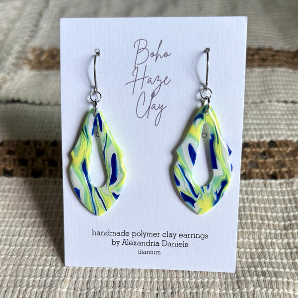 A pair of handmade polymer clay dangle earrings with a blue, yellow, and green marbled pattern, displayed on a white card that reads 'Boho Haze Clay' by Alexandria Daniels.