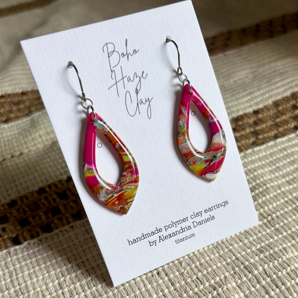 Teardrop-shaped polymer clay dangle earrings with a vibrant pink, orange, yellow, and white marbled pattern, displayed on a white card labeled 'Boho Haze Clay' by Alexandra Daniels.