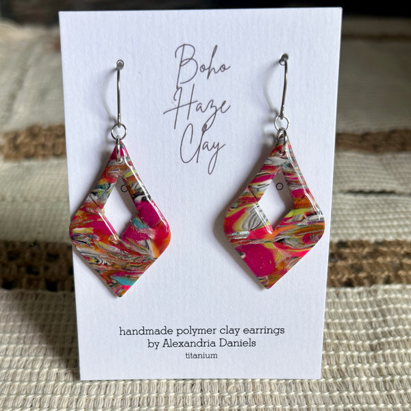A pair of handmade polymer clay dangle earrings by Alexandria Daniels, featuring a vibrant pink, orange, and white marbled pattern, displayed on a white card labeled 'Boho Haze Clay'.