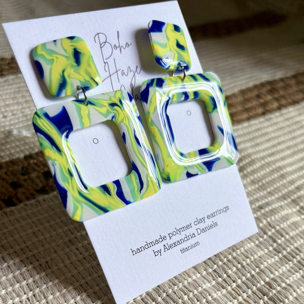 A pair of handmade polymer clay earrings with a blue, green, and yellow marbled pattern, displayed on a white card labeled 'Boho Haze Clay' by Alexandria Daniels.