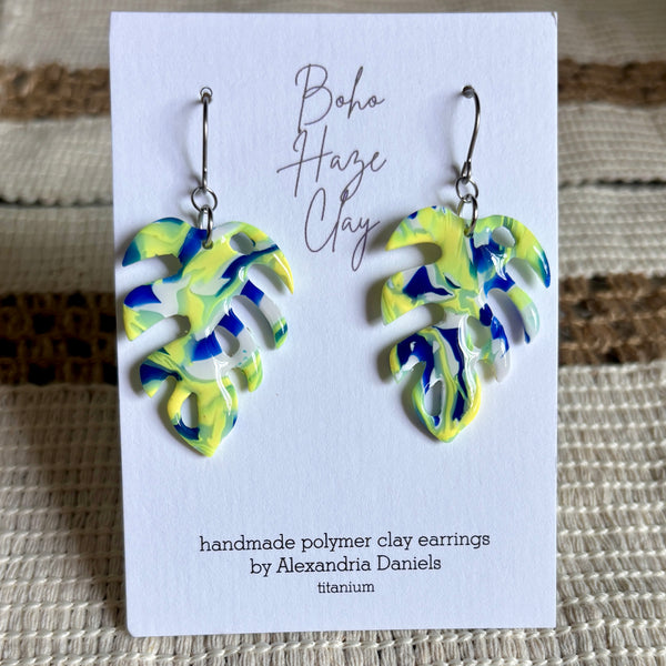 A pair of handmade polymer clay earrings shaped like monstera leaves, featuring a marbled swirl of yellow, blue, and white clay, displayed on a white card with the brand name 'Boho Haze Clay'.