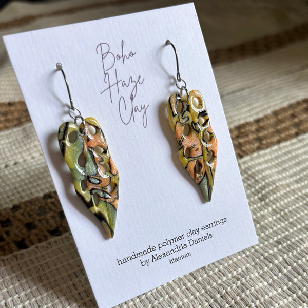A pair of handmade polymer clay dangle earrings displayed on a white card. The earrings feature organic, marbled patterns in olive green, terracotta orange, and black, suspended from silver fishhook wires.