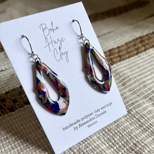 A pair of colorful Boho Haze Clay teardrop earrings displayed on a white card. The earrings feature a marbled pattern of blue, green, red, and white with metallic gold foil accents.