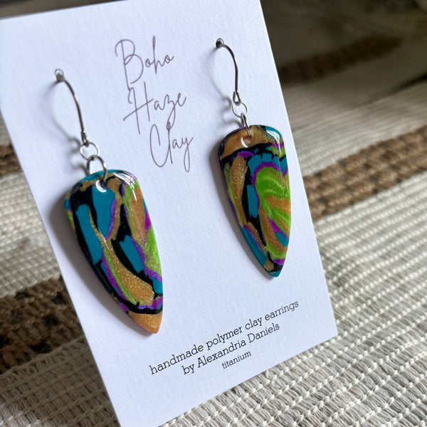 A pair of handmade polymer clay earrings displayed on a white branded card. The earrings have a marbled pattern of teal, black, purple, green, and gold glitter, suspended from silver fishhook wires.