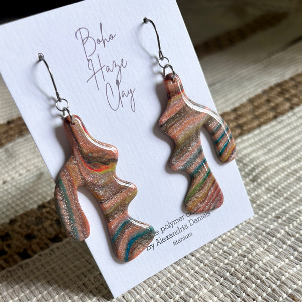 A pair of abstract sculptural dangle earrings made from marbled clay or polymer clay, displayed on a white card labeled 'Boho Haze Clay' by Alexandra Donelly. The earrings feature swirling patterns of terracotta, orange, teal, green, and gold glitter, hanging from silver fishhook wires.