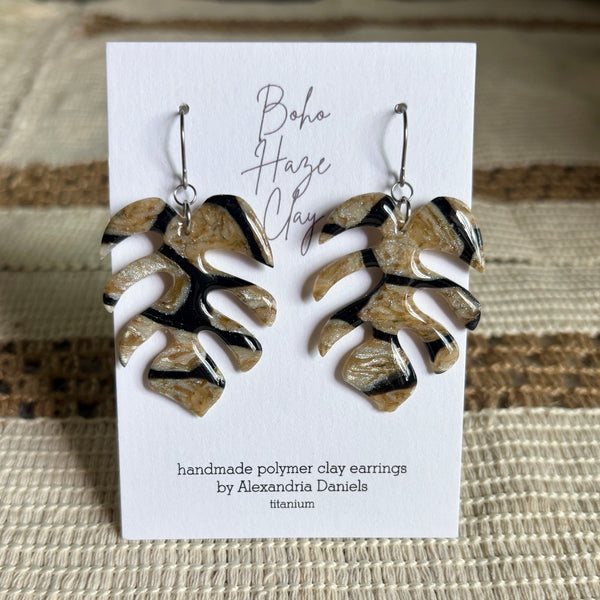 A pair of handmade polymer clay earrings shaped like monstera leaves, displayed on a white card. The earrings feature a metallic titanium base with black and beige swirls.