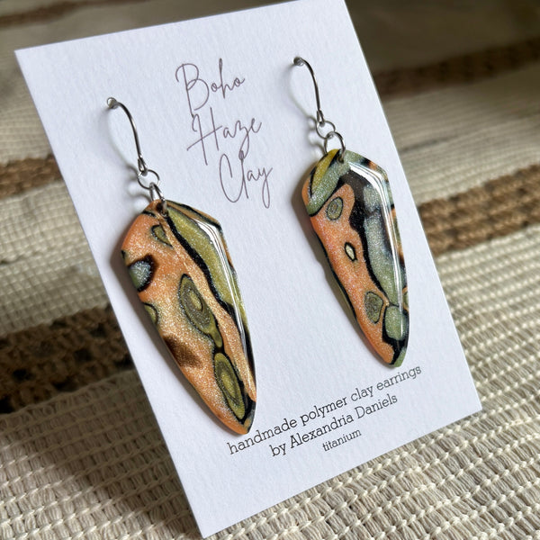A pair of handmade polymer clay dangle earrings displayed on a white branded card. The earrings have an organic, marbled pattern in metallic gold, olive green, black, and a warm peachy-orange color, suspended from silver fishhook wires.