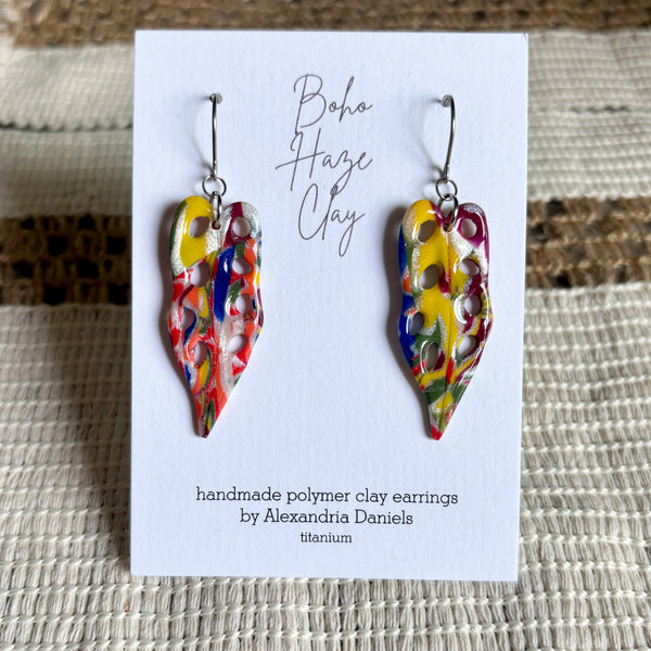 A pair of colorful handmade polymer clay earrings displayed on a white card. The earrings have a teardrop shape with a marbled, abstract pattern of red, yellow, blue, green, and white.