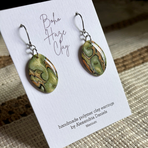 A pair of handmade polymer clay drop earrings with green, gold, and brown marbled patterns, displayed on a white card that reads 'Boho Haze Clay' and 'handmade polymer clay earrings by Alexandra Daniels'.