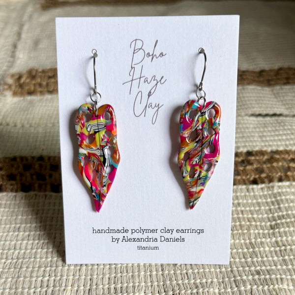 A pair of handmade polymer clay dangle earrings by Boho Haze Clay, displayed on a white card. The earrings are made from a colorful, marbled clay mixture of pink, yellow, orange, and white, suspended from silver fishhook wires.