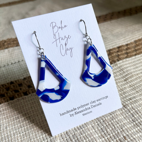 Blue and white marbled polymer clay dangle earrings displayed on a white card that reads 'Boho Haze Clay' and 'handmade polymer clay earrings by Alexandria Daniels titanium'.