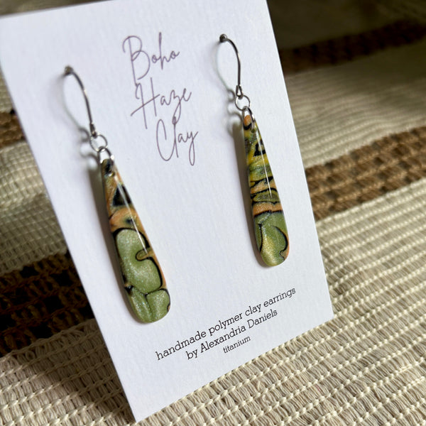 A pair of handmade polymer clay dangle earrings displayed on a white card. The earrings have a marbled pattern of olive green, tan, and black, suspended from silver fishhook wires.