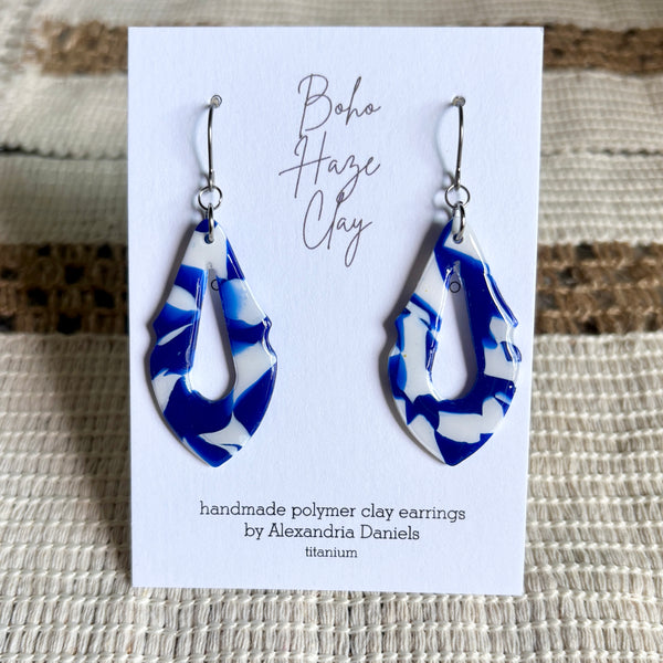 A pair of blue and white marbled polymer clay dangle earrings displayed on a white card that reads 'Boho Haze Clay' and 'handmade polymer clay earrings by Alexandria Daniels'. The card is placed on a textured beige woven surface.