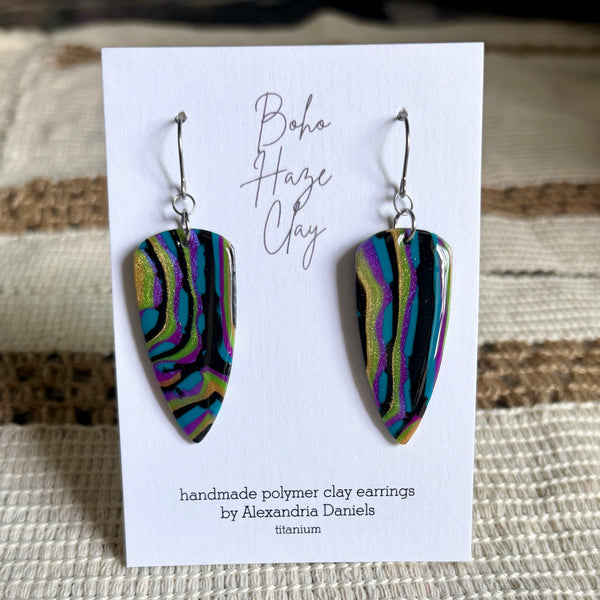 A pair of handmade polymer clay dangle earrings displayed on a white card. The earrings have a glossy, marbled finish with swirls of black, purple, green, and gold, suspended from silver fishhook wires.