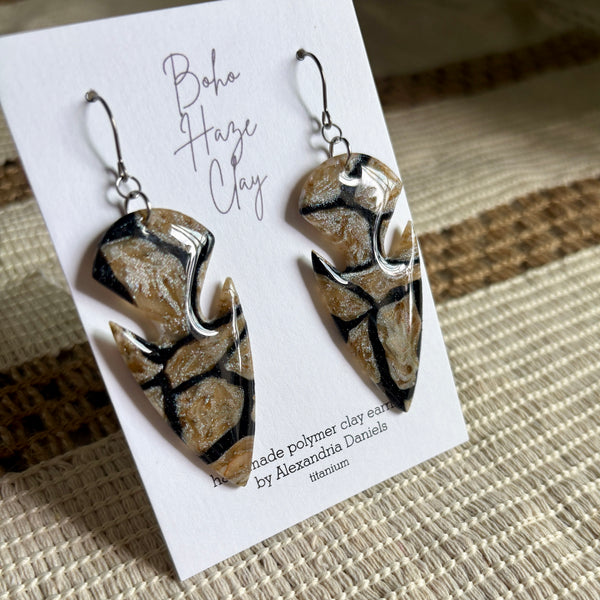 A pair of handmade polymer clay dangle earrings displayed on a white card labeled 'Boho Haze Clay' by Alexandra Daniela, resting on a woven beige and brown surface.