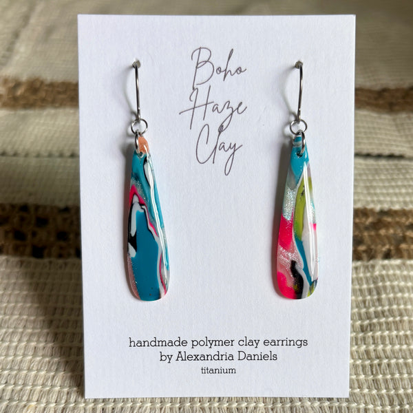 A pair of handmade polymer clay dangle earrings with a colorful marbled pattern in turquoise, pink, white, and black, displayed on a white card that reads 'Boho Haze Clay' by Alexandria Daniels.