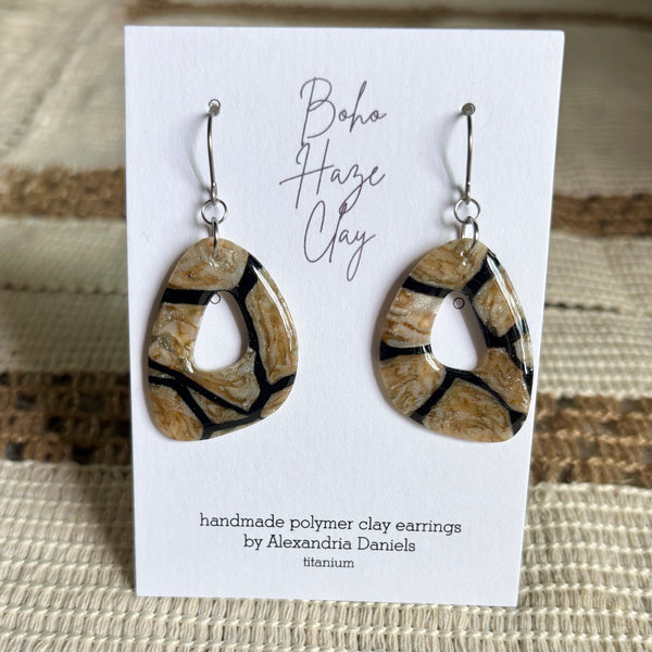 A pair of handmade teardrop-shaped polymer clay earrings with a brown, beige, and black marbled pattern, displayed on a white card labeled 'Boho Haze Clay' by Alexandria Daniels.