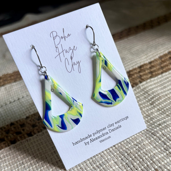 A pair of handmade teardrop-shaped polymer clay earrings with a blue, green, and white marbled pattern, displayed on a white card that reads 'Boho Haze Clay' by Alexandra Daniels, set against a woven beige background.