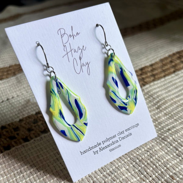 A pair of handmade polymer clay dangle earrings with a marbled swirl pattern of blue, lime green, and yellow, displayed on a white card that reads 'Boho Haze Clay' by Alexandria Daniels, set against a woven beige background.