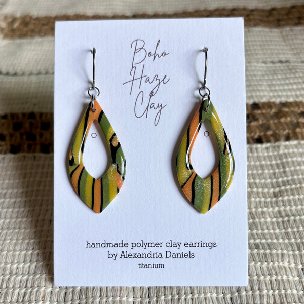Teardrop-shaped polymer clay earrings with a green, orange, and black striped pattern, displayed on a white card labeled 'Boho Haze Clay' by Alexandria Daniels.