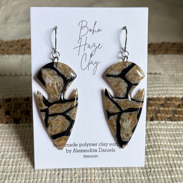 Handmade polymer clay dangle earrings by Alexandria Daniels, displayed on a white backing card. The earrings are made from beige colored clay with black and silver veining, designed in an arrowhead shape.