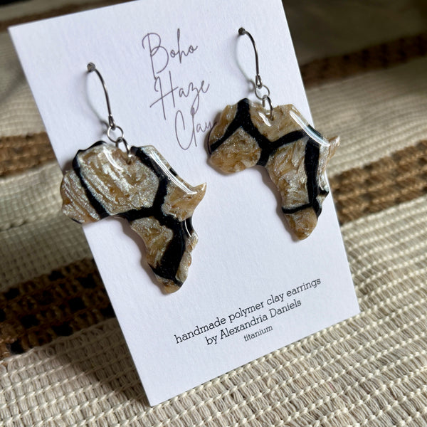 A pair of handmade polymer clay earrings shaped like the African continent, featuring a textured beige and brown base with black veins, displayed on a white card labeled 'Boho Haze Clay' by Alexandria Daniels.