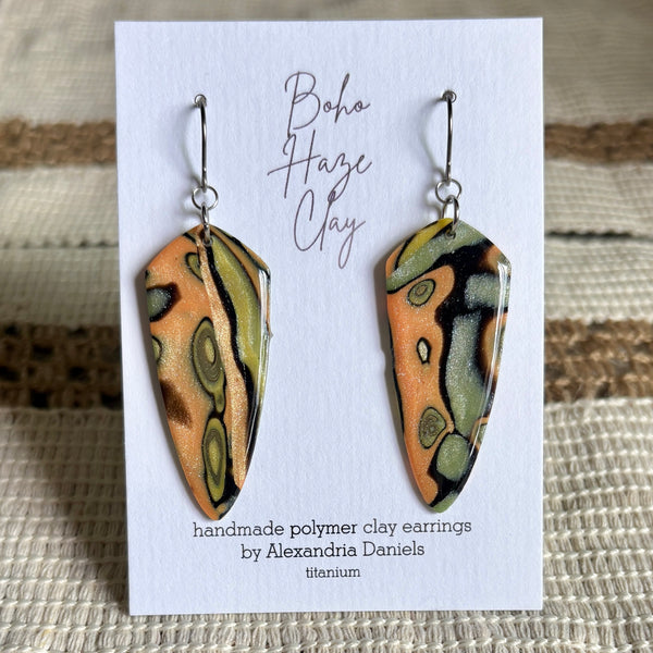 A pair of handmade polymer clay dangle earrings by Alexandria Daniels, displayed on a white card. The earrings feature a metallic orange, olive green, and black marbled pattern on a titanium base.