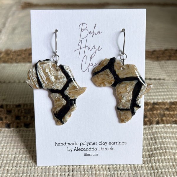 Two handmade polymer clay earrings shaped like the continent of Africa featuring a textured beige and gold base with bold black lines, displayed on a white card that reads 'Boho Haze Clay' by Alexandria Daniels.