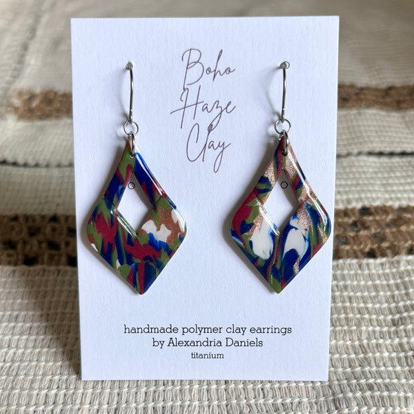 A pair of handmade polymer clay dangle earrings displayed on a white card. The earrings have a marbled pattern of blue, red, green, and white, suspended from silver fishhook wires.