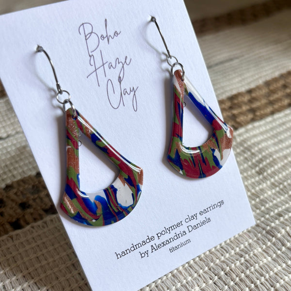 A pair of colorful teardrop-shaped polymer clay earrings displayed on a white card that reads 'Boho Haze Clay' and 'handmade polymer clay earrings by Alexandria Daniels titanium.' The earrings have a marbled pattern of blue, red, green, and white with metallic gold flecks, hanging from silver fishhook wires.
