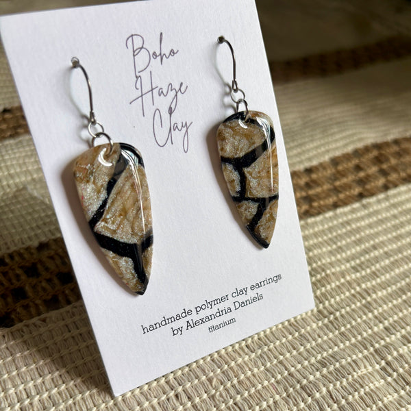 A pair of handmade polymer clay earrings displayed on a white card. The earrings are teardrop-shaped with a marbled pattern of beige, brown, and black, hanging from silver fishhook wires.