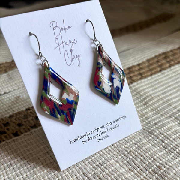 A pair of dangling geometric earrings displayed on a white card. The earrings have a marbled pattern of blue, red, green, and white, with silver fishhook wires.