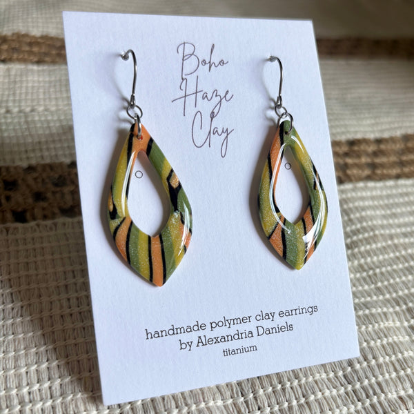 Teardrop-shaped polymer clay earrings with a green, orange, and black marbled pattern, displayed on a white card labeled 'Boho Haze Clay' by Alexandria Daniels.
