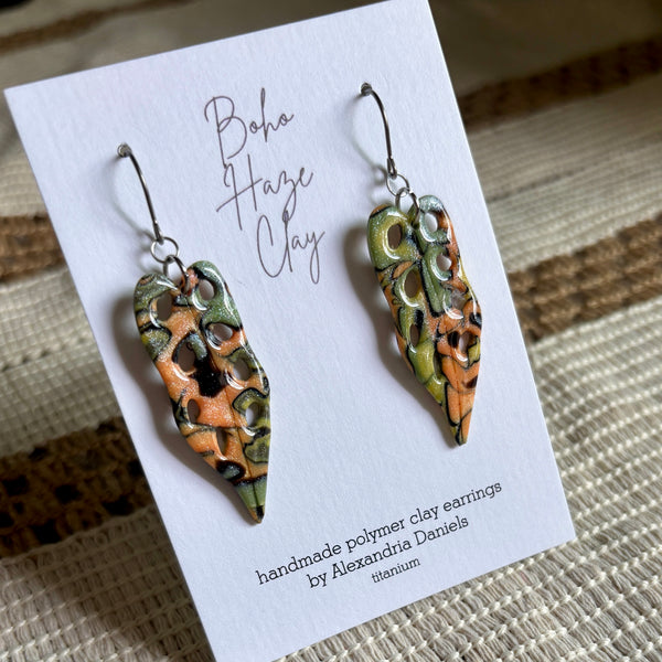 A pair of handmade polymer clay earrings by Alexandria Daniels, displayed on a white card. The earrings feature a textured, organic pattern in earthy tones of orange, olive green, and black, suspended from silver fishhook wires.