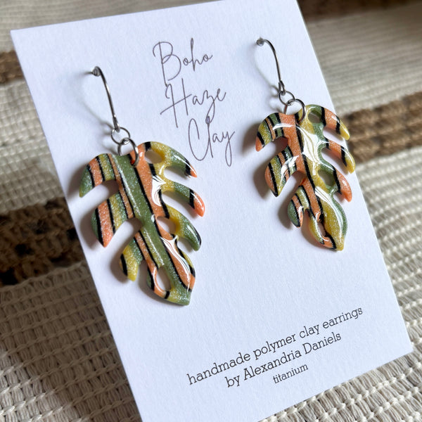 A pair of handmade polymer clay earrings shaped like tropical monstera leaves, displayed on a white card. The earrings feature a marbled pattern of orange, olive green, black, and cream.