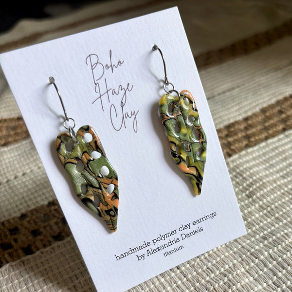 A pair of handmade polymer clay earrings displayed on a white card. The earrings have a marbled pattern of olive green, orange, and black with small white inclusions, suspended from silver fishhook wires.