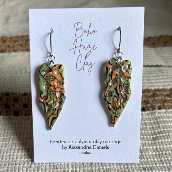 A pair of handmade polymer clay dangle earrings by Alexandria Daniels, displayed on a white card. The earrings feature organic, marbled patterns of olive green, terracotta orange, and metallic silver.