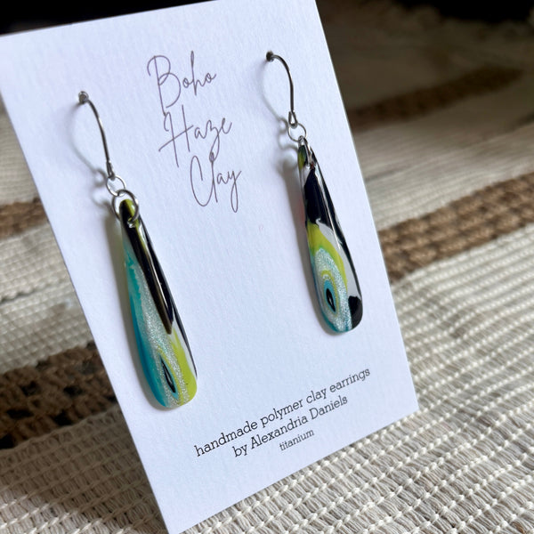 A pair of handmade polymer clay dangle earrings displayed on a white card. The earrings feature a teardrop shape with a marbled swirl pattern of black, teal, lime green, and white, suspended from silver fishhook wires.