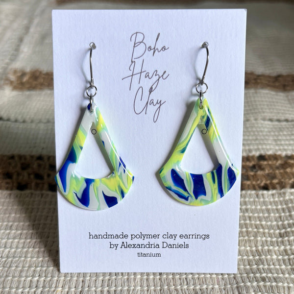 A pair of handmade polymer clay dangle earrings with a blue, green, and white marbled pattern, displayed on a white card labeled 'Boho Haze Clay' by Alexandria Daniels.