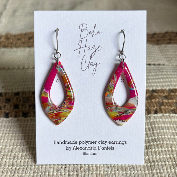 A pair of handmade polymer clay dangle earrings with a bright pink, orange, yellow, and white marbled pattern, displayed on a white card labeled 'Boho Haze Clay' by Alexandria Daniels.