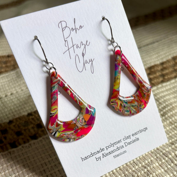 A pair of colorful teardrop-shaped polymer clay earrings displayed on a white branded card. The earrings feature a vibrant marbled pattern with pink, orange, yellow, green, and blue.