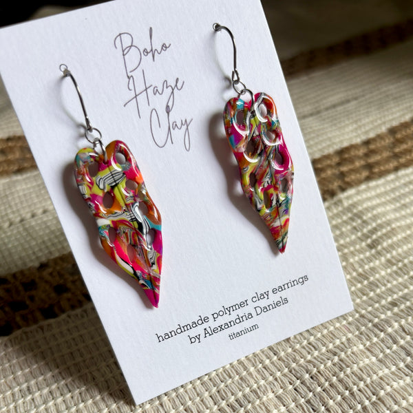 A pair of colorful handmade polymer clay dangle earrings displayed on a white card labeled 'Boho Haze Clay' by Alexandria Daniels, placed on a woven beige surface.