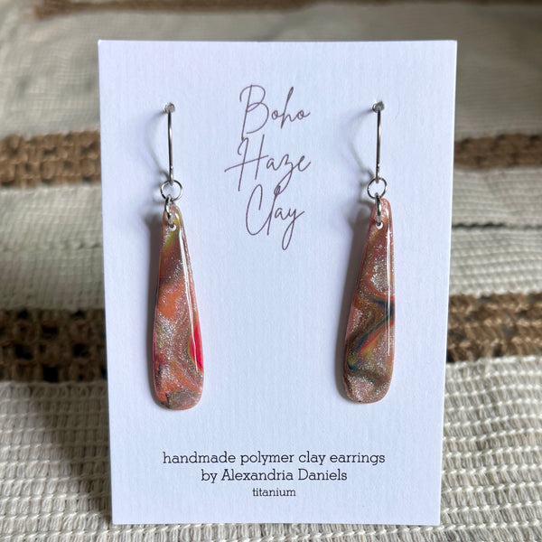 A pair of handmade polymer clay dangle earrings by Alexandria Daniels, displayed on a white card. The earrings are made from a metallic titanium-colored clay with a marbled pattern of orange, red, and green, hanging from silver fishhook wires.