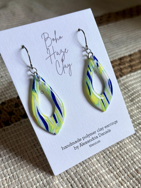 A pair of handmade polymer clay dangle earrings by Boha Haze Clay, displayed on a white card. The earrings feature a marbled pattern of blue, lime green, and white, suspended from silver fishhook wires.