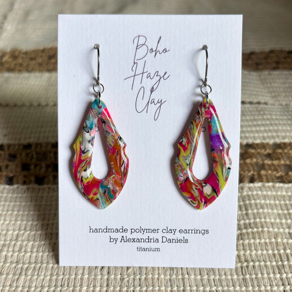 A pair of colorful handmade polymer clay dangle earrings displayed on a white card. The earrings have an abstract marbled pattern with pink, yellow, green, blue, and white swirls, hanging from silver-toned fishhook wires.