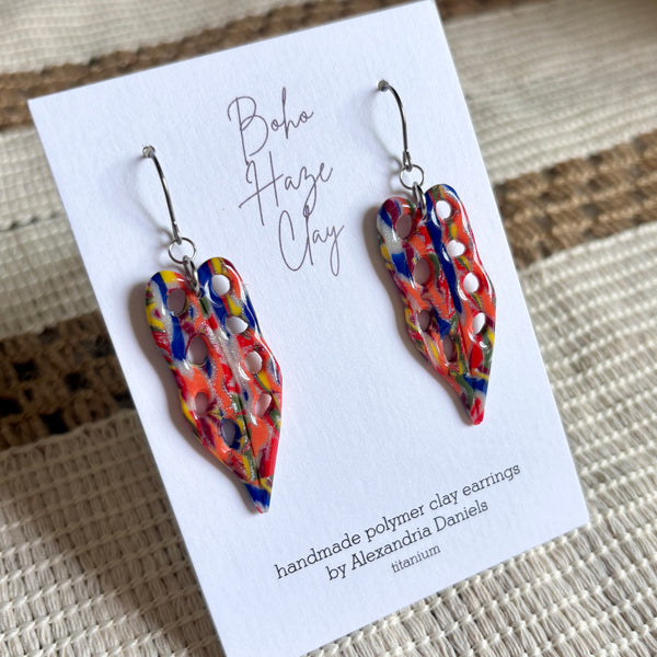 A pair of colorful handmade polymer clay earrings displayed on a white card. The earrings have an abstract, marbled pattern with red, orange, yellow, blue, and white swirls, hanging from silver fishhook wires.