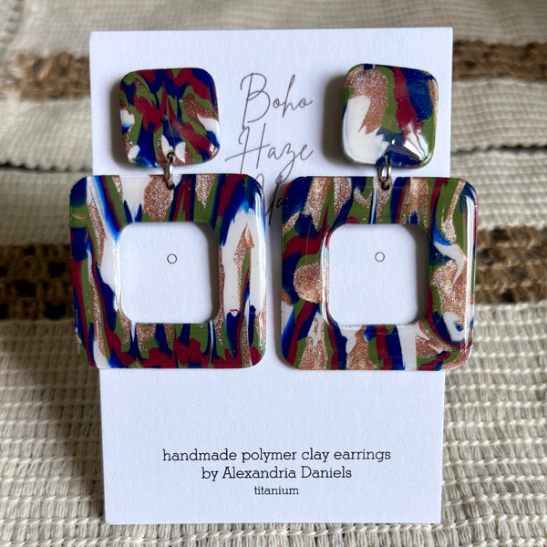 A pair of handmade polymer clay earrings by Alexandria Daniels, featuring a square drop design with a smaller multicolor top piece and a larger square bottom piece. The earrings have a marbled pattern of blue, red, green, white, and metallic gold glitter.