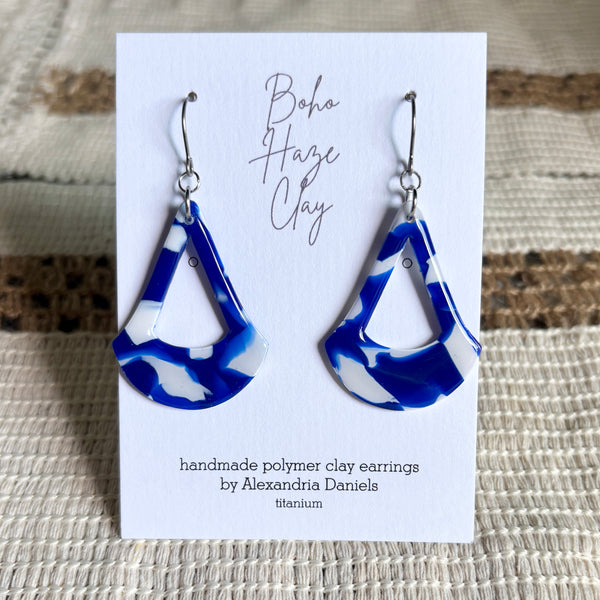 Blue and white marbled polymer clay dangle earrings displayed on a white card that reads 'Boho Haze Clay' and 'handmade polymer clay earrings by Alexandria Daniels', set against a woven beige fabric background.