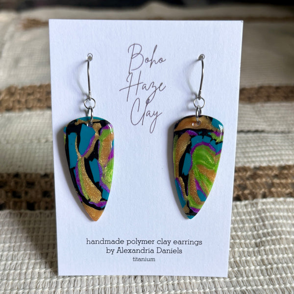 A pair of handmade polymer clay dangle earrings displayed on a white card. The earrings have an abstract marbled pattern with black, turquoise, green, purple, and gold glitter, suspended from silver fishhook wires.