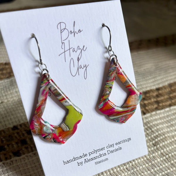 A pair of colorful Boho Haze Clay geometric drop earrings displayed on a white card. The earrings feature a marbled, swirling pattern of pink, orange, yellow, and white, suspended from silver fishhook wires.